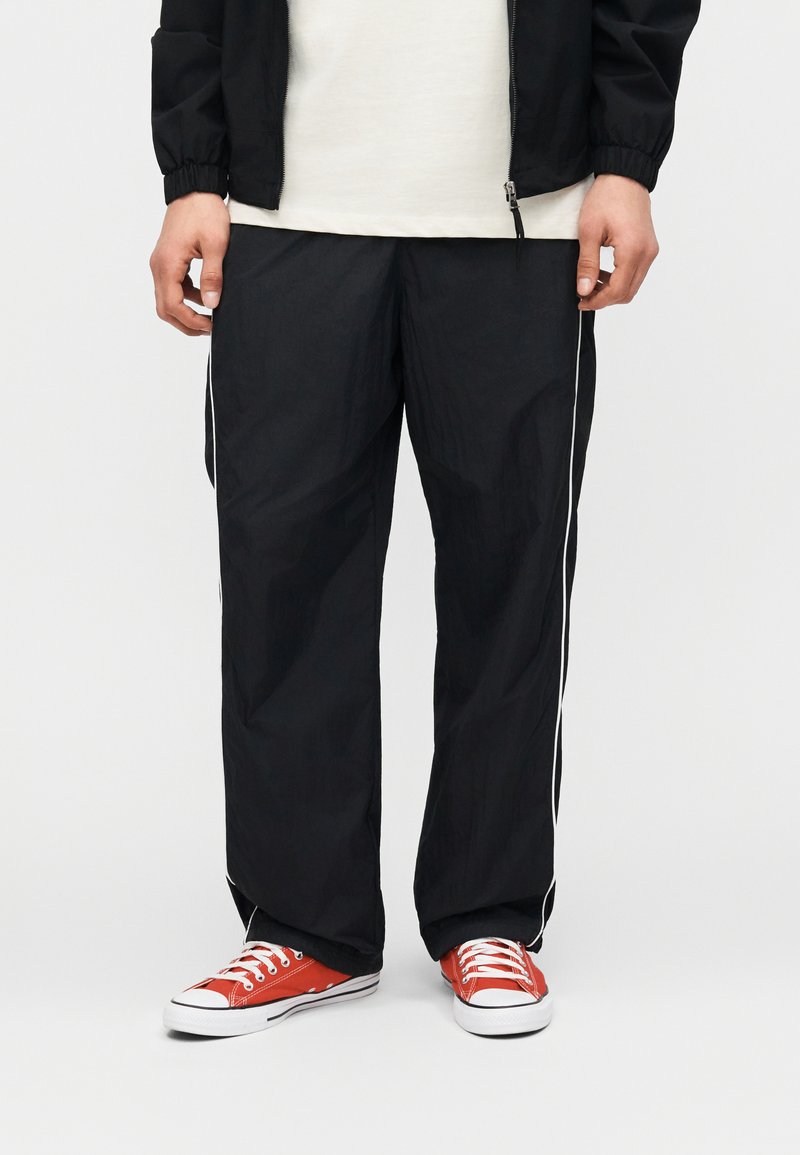 Άτομο που φοράει μαύρο φόρμα με λευκές λωρίδες στα πλάγια, κόκκινα αθλητικά παπούτσια χαμηλού προφίλ και μαύρο τζάκετ με φερμουάρ πάνω από λευκό πουκάμισο.