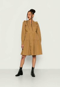 Vestido de cor camelo, com manga longa, destacando um colarinho rufado, fecho de botão e barra em camadas com padrões texturizados subtis. Botas de tornozelo pretas.