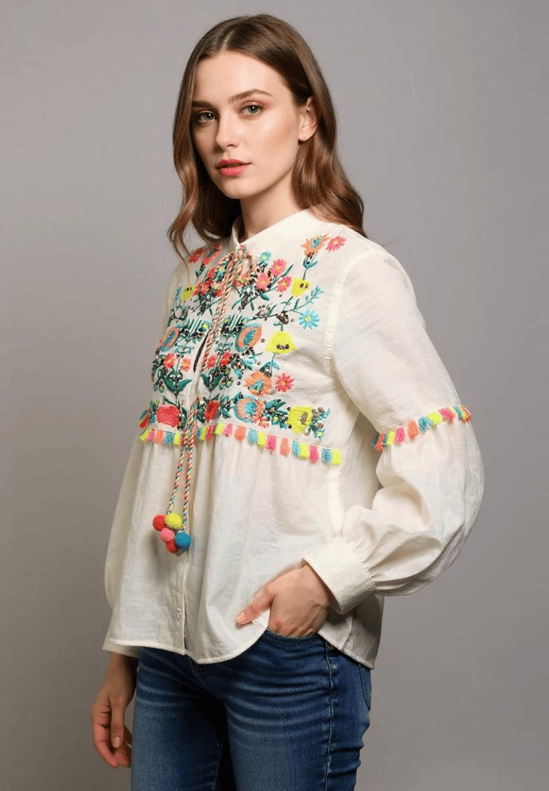 Blusa de algodón blanca con colorido bordado floral, acentos de borlas y un cordón con pompones. Mangas largas con volumen.