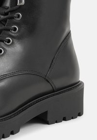 Bottine en cuir noir avec design à lacets, œillets métalliques, semelle noire robuste et coutures visibles sur le côté.