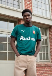 Teal sportströja med svart krage, logotyp och sponsorskrift. Modellen bär beiga byxor och står framför en murverksbyggnad.