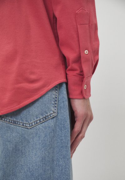 Růžové dlouhé rukávy košile z texturované látky s knoflíkovými manžetami, kombinované se světle modrými džínovými kalhotami s prošívanou kapsou.