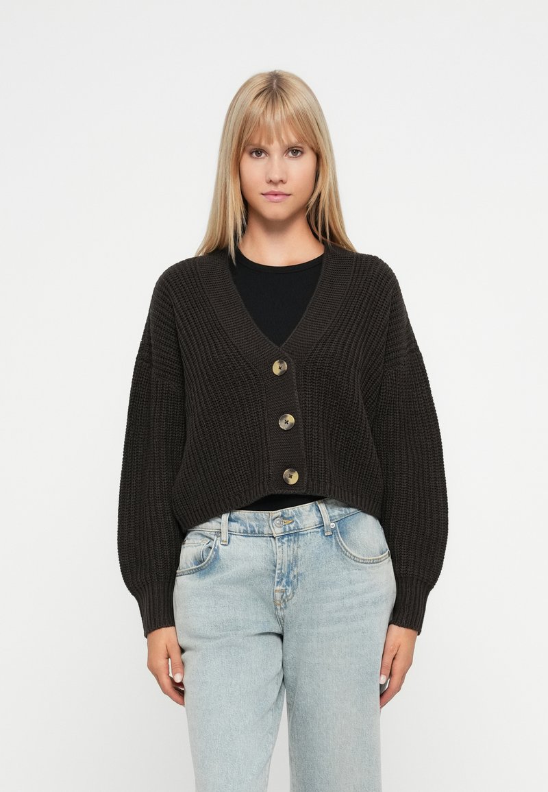 Temno rjava pletena cardigan jakna z V-izrezom, sprednjim gumbom in širokimi rokavi. Oblečena čez črn top in svetlo modre kavbojke.
