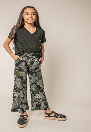 Jeune fille debout, les mains dans les poches, portant un t-shirt noir moucheté, un pantalon large à motifs et des sandales noires.