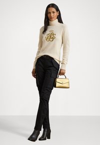 Femeie îmbrăcată cu pulover crem cu guler înalt, având un monogram de aur, pantaloni cargo negri, cizme negre cu toc, ținând o mică geantă de umăr aurie.