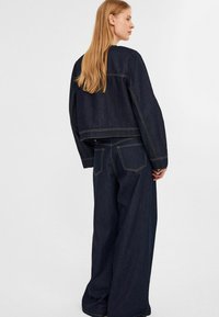 Mörk denimjacka med vida ärmar och en kort design, kombinerad med matchande högmidjade, vida jeans. Gula sömmar som accent.