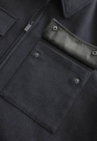 Camicia di lana blu scuro con chiusura a zip, due tasche piatte e un tessuto testurizzato. Dettagli in nylon nero su una tasca con bottoni automatici.