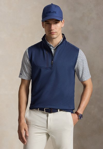 Homme portant une casquette bleu marine, un gilet zippé bleu marine par-dessus une chemise à manches courtes à motifs, un pantalon beige et une montre avec un bracelet en cuir marron.