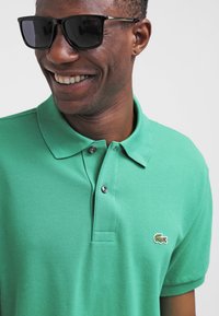 Grön polo skjorta i texturerad bomull, med klassisk krage, tvåknäppning och en liten Lacoste-logotyp på vänster bröst.