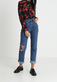 Sort blomsterbluse med blonde detaljer, højtaljede blå denimjeans med huller og sorte snørede sko, sat mod en ensfarvet baggrund.