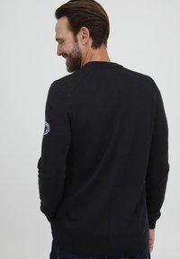 Pulover tricotat de culoare bleumarin, cu accente de piele neagră pe umeri, manșete și tiv cu aspect ondulat. Prezintă un guler rotund și un mic patch cu logo pe mânecă.