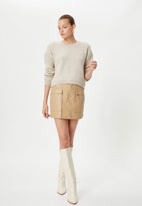 Beżowy sweter o prążkowanym wzorze, brązowa skórzana mini spódnica z dwiema przednimi kieszeniami oraz wysokie białe botki. Prosty, elegancki design z gładkimi teksturami.
