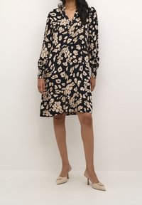 Schwarzes Kleid mit floralem Muster in Creme und Beige, lange Ärmel, V-Ausschnitt und knieumspielender Schnitt, strukturierten Stoff. Getragen mit hellen Schuhen.