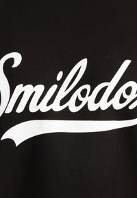 Svart tyg som visar ordet "Smilodoz" i fet, vit skrift med en böjd understrykning. Slät textur och tydlig typografi.