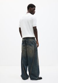 Hvid kortærmet strikket top og løse blå denimjeans med brede ben, fremtrædende syninger og store baglommer, set fra bagsiden.
