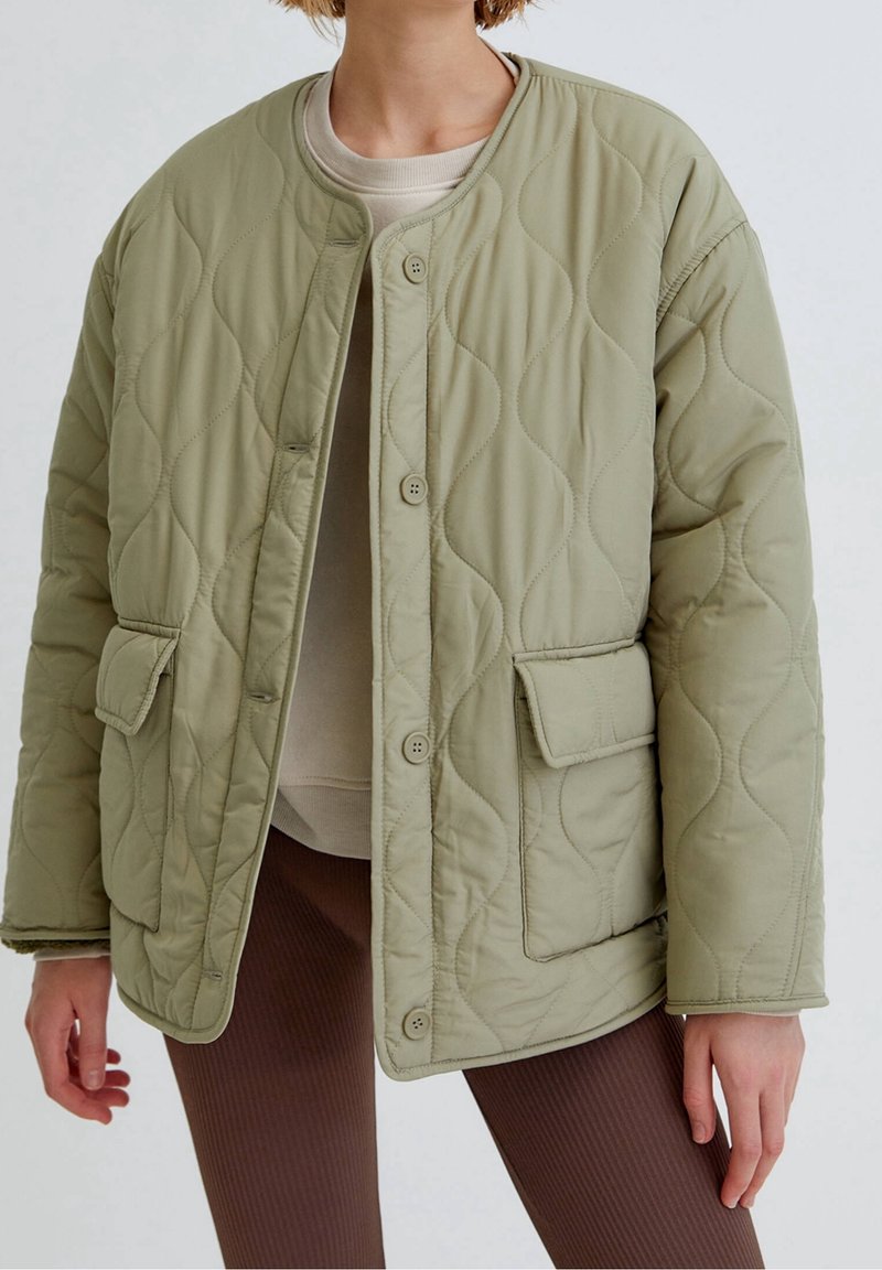 Personne portant une veste matelassée vert clair avec boutons et poches avant à rabat, par-dessus un haut beige et un pantalon côtelé marron.