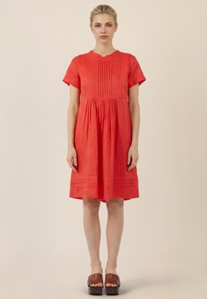 Woman stands facing forward wearing knee-length red dress with short sleeves, pleated details, and brown platform sandals against beige background.