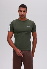 Camiseta ajustada de color verde oliva con mangas cortas, que presenta rayas blancas en los lados y un pequeño parche de logo en el pecho. Textura suave.