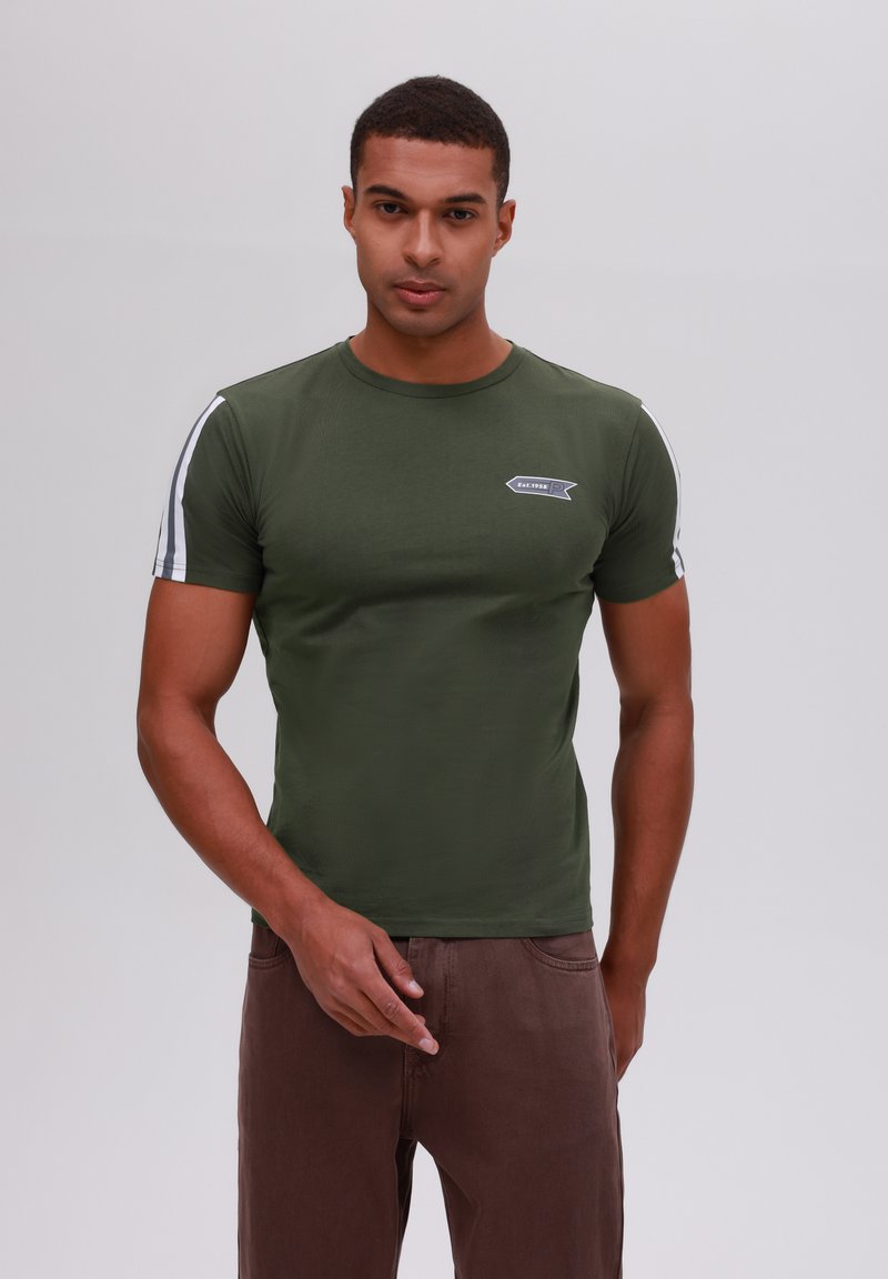 Camiseta ajustada de color verde oliva con mangas cortas, que presenta rayas blancas en los lados y un pequeño parche de logo en el pecho. Textura suave.