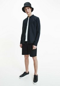 Camicia nera a maniche lunghe, maglietta bianca, pantaloni corti neri, cappello a secchiello nero e sneakers nere con suole bianche. Design semplice e minimalista, senza motivi.
