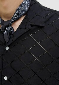 Schwarzes Hemd mit einem Diamantschnitt-Muster und weicher Textur. Weiße Knöpfe und ein dazu passender schwarz-weißer Paisleymuster-Schal unterstreichen den Look.