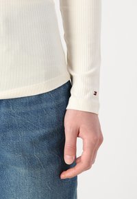Ribbete kremfarget langermet topp med en tettsittende design, med en liten logoakcent på mansjetten, kombinert med blå denimjeans.