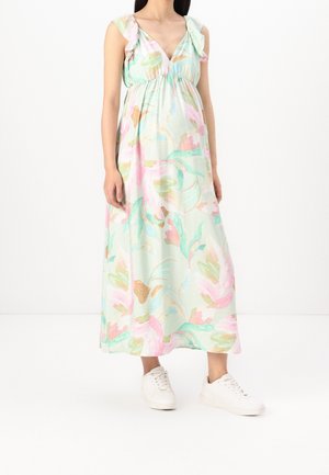 Femme portant une robe maxi fleurie pastel avec des épaules à volants et des baskets blanches, debout devant un fond blanc uni.