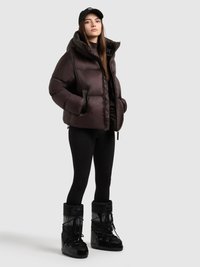 Puffer jacket in dark brown with a high collar, zip closure, and two front pockets. Black leggings and glossy black snow boots complete the look.
