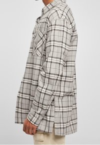 Uomo che indossa una camicia a maniche lunghe a quadri beige e marrone con taschino frontale, abbinata a pantaloni beige chiaro, in piedi di lato su uno sfondo bianco.