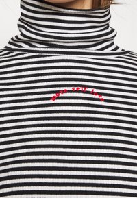 Col roulé rayé noir et blanc avec une texture côtelée et un texte brodé rouge "plus d'amour de soi" sur la poitrine.