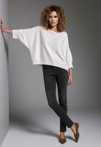 Übergroßer weißer Strickpullover, dunkelblaue enge Jeans, olivgrüne flache Schuhe mit spitz zulaufender Spitze. Modell posiert mit einer Hand an der Wand, neutraler Hintergrund.