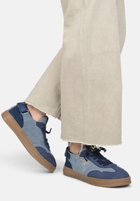 Baskets bleues et grises en suède et tissu, avec un bout rond, une semelle en caoutchouc et des lacets noirs, sur fond de pantalon beige.