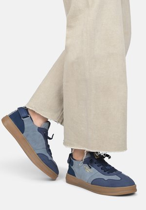 Sneaker blu e grigi con materiali in suede e tessuto, dotati di punta rotonda, suola in gomma e lacci neri, posizionati su pantaloni beige.