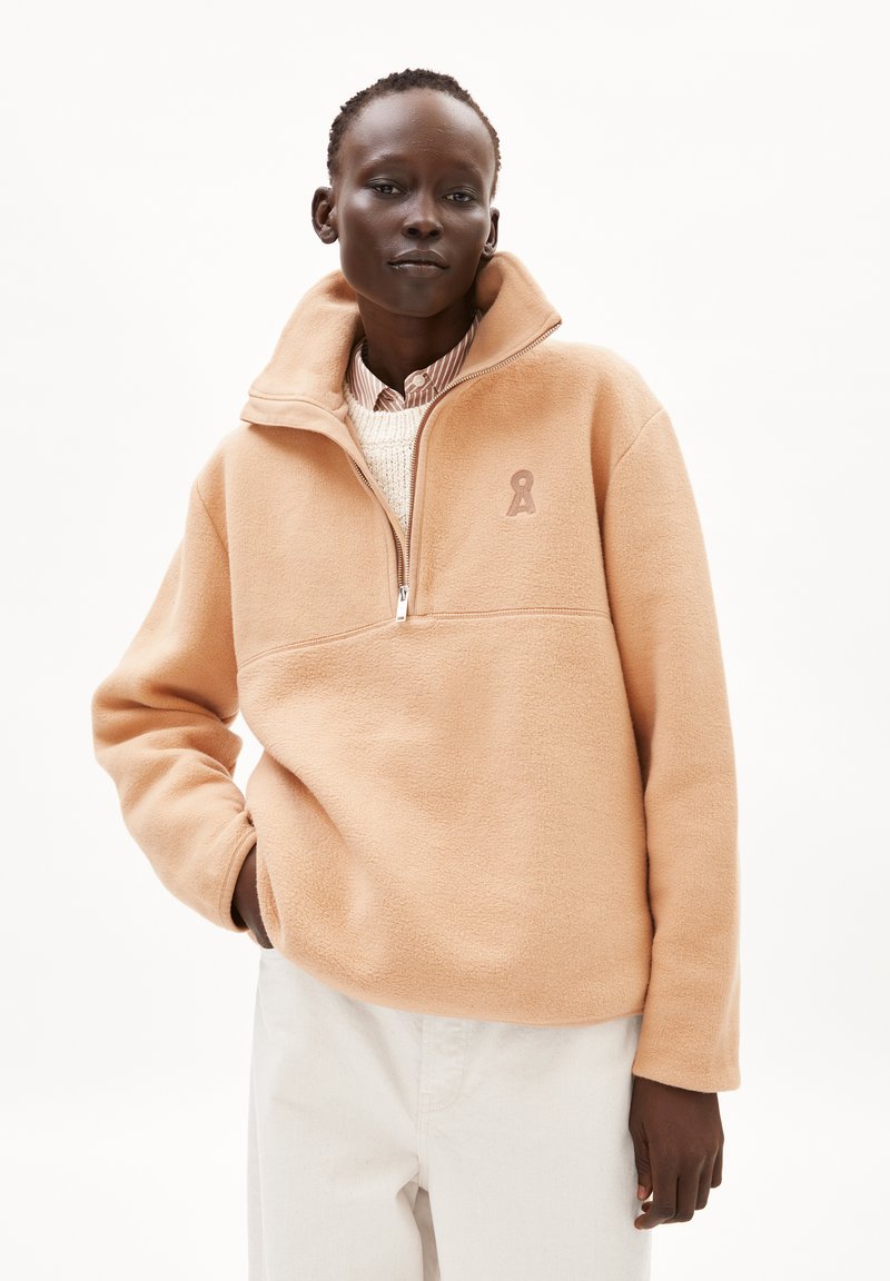 Pfirsichfarbener Fleece-Pullover mit einem Reißverschlusskragen, der ein gesticktes Logo auf der Brust und eine weiche, strukturierte Oberfläche aufweist. Getragen über einem gestreiften Hemd.