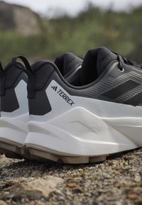 Ασπρόμαυρα παπούτσια Adidas Terrex για τρέξιμο σε μονοπάτι με ταινίες έλξης σε βραχώδη εξωτερική επιφάνεια.