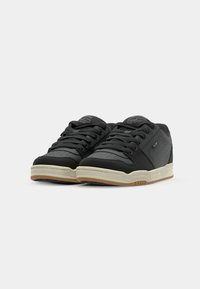 Black athletic shoes with a smooth synthetic upper, perforated detailing, and a cream rubber sole. Features black laces and logo accents.