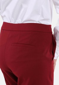 Pantaloni color bordeaux con vita piatta, una tasca posteriore singola e texture liscia. Indossati con una camicia bianca, che evidenzia una silhouette aderente.