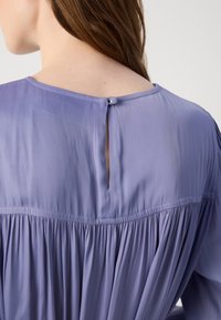 Blusa lavanda con un design plissettato, texture morbida, scollatura rotonda e apertura a occhio di chiave con bottone al retro.