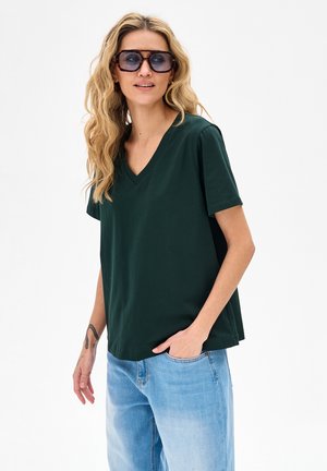 Kobieta o długich blond włosach, nosząca dużą, ciemną okularów przeciwsłonecznych, ciemnozieloną koszulkę z dekoltem w serek, jasnoniebieskie dżinsy oraz widoczne tatuaże na ramieniu.