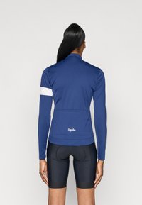 Jersey de ciclismo de manga longa azul com uma risca branca no braço e bolsos nas costas, combinado com calções de ciclismo pretos. Textura de tecido suave.