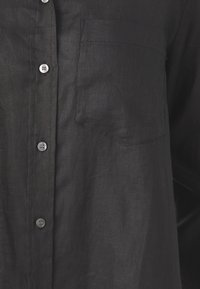 Chemise en lin noire avec une patte de boutonnage à l'avant, des boutons argentés, une poche poitrine unique et une finition douce et texturée.