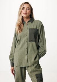 Camicia verde oliva con bottoni, taschino sul petto, tessuto testurizzato e maniche lunghe con accenti di verde scuro sui polsini.