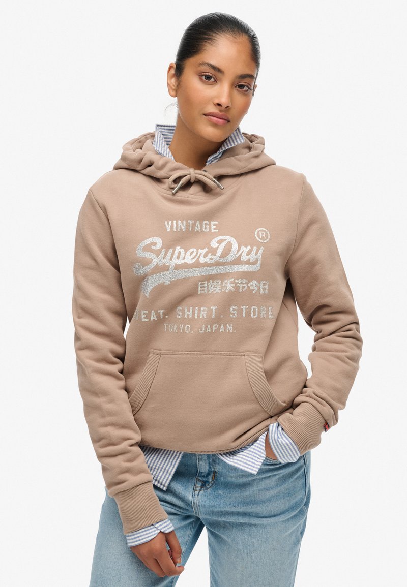Hoodie marron avec capuche à cordon de serrage, poche avant et texte imprimé en argent. Superposé avec une chemise rayée bleue et blanche en dessous.