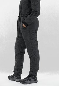 Zwart gewatteerd jumpsuit met een trekkoord in de taille, zijzakken en ribbelmanchetten, gecombineerd met stevige zwarte wandelschoenen.