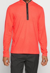 Homme portant un pull-over long à manches longues corail vif avec une fermeture éclair noire au quart, accompagné d'un pantalon gris foncé, se tenant devant un fond uni.