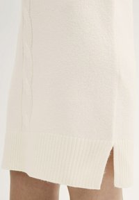 Strickkleid in Creme mit geripptem Saum, strukturiertem Oberflächenmaterial und seitlichen Schlitzen. Mit dezenten Zopfmuster-Details entlang der Seite.