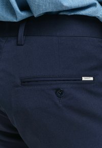 Marineblå bukser med teksturert stoff, med en enkelt baklomme med knapp og en liten hvit merkelapp med merkevarenavnet "GANT."