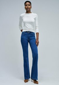 Femme debout portant un haut blanc à manches longues, un jean bleu taille haute avec une ceinture, et des chaussures à talons aiguilles beiges.