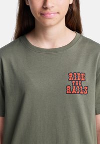 Camiseta de algodón color verde oliva con cuello redondo y mangas cortas; presenta un gráfico en rojo y negro que dice "RIDE THE RAILS" en el lado izquierdo del pecho.