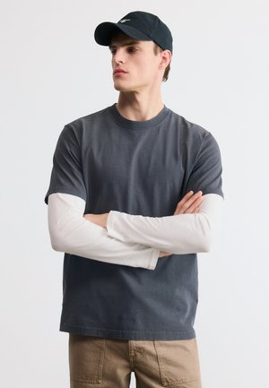 Νέος άνδρας με σταυρωμένα χέρια, φορεί μαύρο καπέλο, σκούρο γκρι κοντομάνικη μπλούζα πάνω από λευκά μακριά μανίκια και μπεζ παντελόνι, πάνω σε λευκό φόντο.